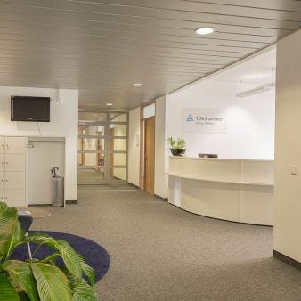 Empfangsbereich im B&uuml;ro mit Rezeptionstresen, T&Uuml;V Rheinland-Logo an der Wand, Schlie&szlig;f&auml;chern und Pflanzen vor einem Durchgang zu weiteren R&auml;umen.