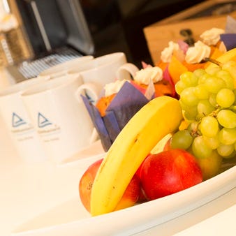 Schale mit Banane, Trauben und &Auml;pfeln neben kleinen Muffins und Kaffeetassen mit T&Uuml;V Rheinland-Logo auf wei&szlig;em Tablett.