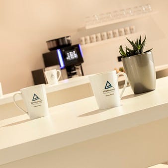 Drei wei&szlig;e Kaffeetassen mit T&Uuml;V Rheinland-Logo auf einem Tresen, im Hintergrund Kaffeemaschine und kleine Topfpflanze.