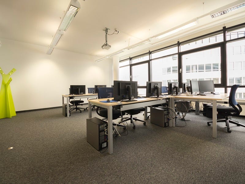 Seminarraum mit mehreren PC-Arbeitspl&auml;tzen, Desktop-PCs und Monitoren, Deckenprojektor, gro&szlig;en Fenstern und gr&uuml;ner Garderobe.