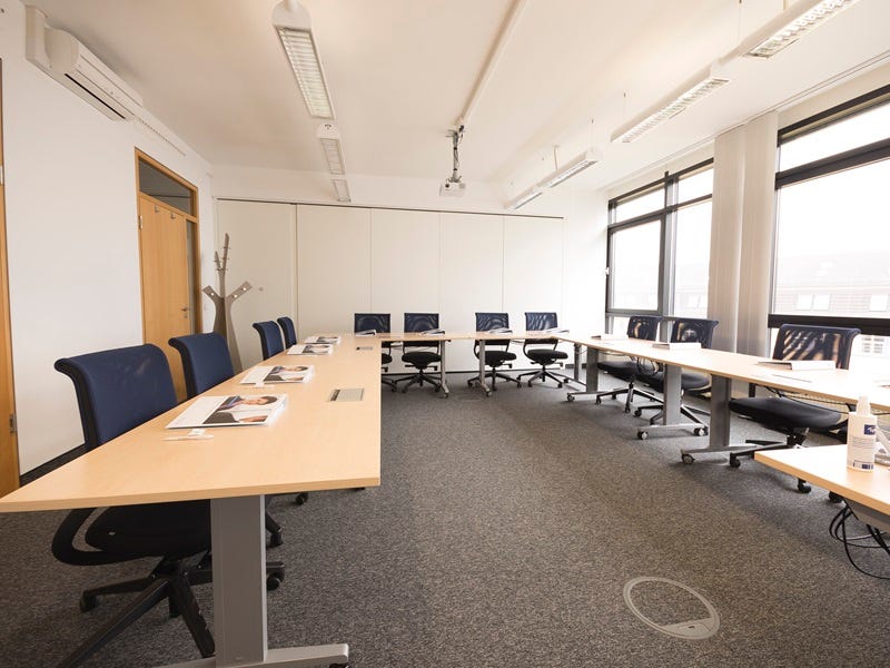 U-f&ouml;rmige Bestuhlung in einem hellen Schulungsraum mit blauen St&uuml;hlen, Deckenprojektor und gro&szlig;en Fenstern.