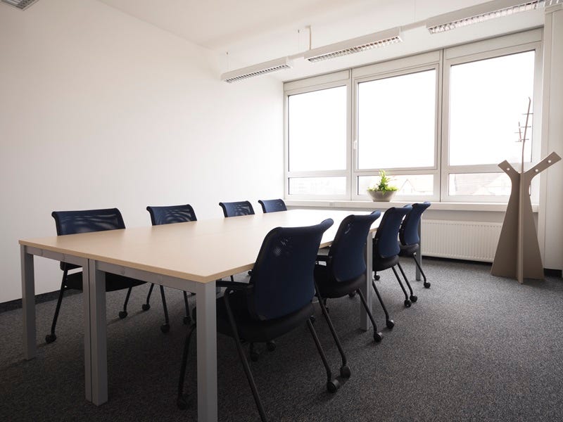 Heller Besprechungsraum mit rechteckigem Tisch, acht blauen St&uuml;hlen, gro&szlig;em Fenster, Pflanze und Garderobenst&auml;nder.