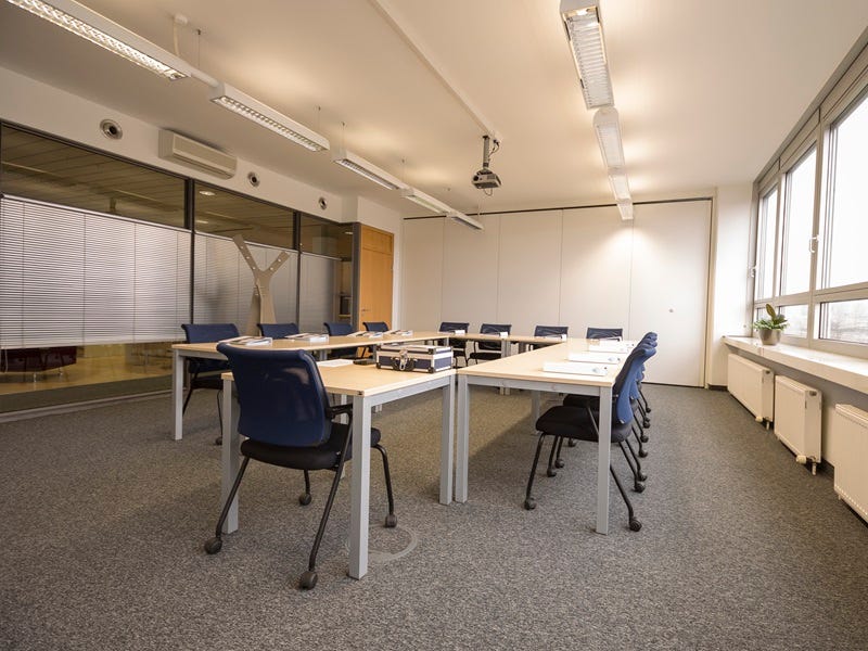 Heller Schulungsraum mit U-f&ouml;rmigen Tischen, blauen St&uuml;hlen, Deckenprojektor und gro&szlig;en Fenstern f&uuml;r Seminare und Meetings.