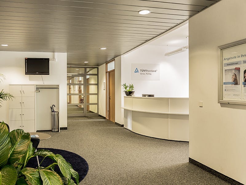 Das Bild zeigt einen modernen Empfangsbereich der TÜV Rheinland Akademie Stuttgart. Im Zentrum des Bildes steht ein geschwungener Empfangstresen, der schlicht und einladend gestaltet ist.