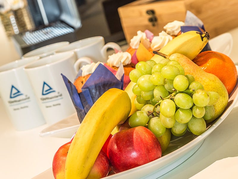 Das Bild zeigt ein Arrangement mit frischen Früchten und Gebäck auf einem Tisch. Im Vordergrund befinden sich eine Schale mit verschiedenem Obst, darunter grüne Trauben, eine Banane, rote Äpfel und eine Birne. 
