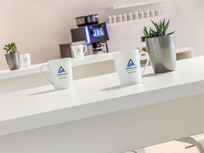 Drei wei&szlig;e Kaffeetassen mit T&Uuml;V Rheinland-Logo auf einem Tresen, im Hintergrund Kaffeemaschine und kleine Topfpflanze.