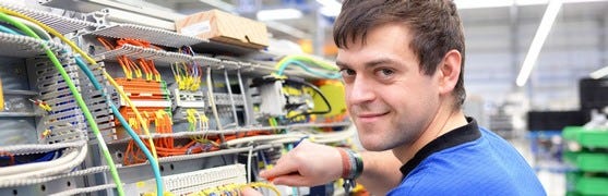 Ein Techniker arbeitet an einer Schalttafel mit farbigen Kabeln und Leitungen ausgestattet, die sorgfältig geordnet und angeschlossen sind. Im Hintergrund sind weitere industrielle Geräte und Vorrichtungen zu sehen.