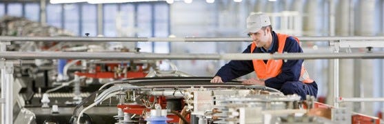 Das Bild zeigt einen Techniker in einer Fabrikhalle, der an einer großen industriellen Anlage arbeitet. Er trägt eine Sicherheitsweste und einen Helm und überprüft akribisch die Komponenten der Maschine.