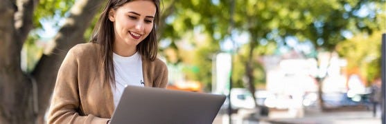Junge Frau sitzt draußen vor einem Baum und arbeitet an einem Laptop 