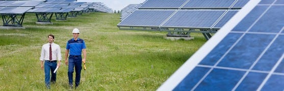 Das Bild zeigt zwei Männer, die durch ein Feld mit zahlreichen Solarpaneelen gehen. Einer trägt einen Anzug, der andere Arbeitskleidung und einen Helm. Das Bild vermittelt den Eindruck von technischer Inspektion und nachhaltiger Energiegewinnung.