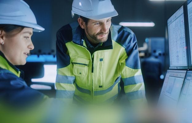 Eine Frau und ein Mann mit Sicherheitskleidung schauen auf verschiedene Bildschirme