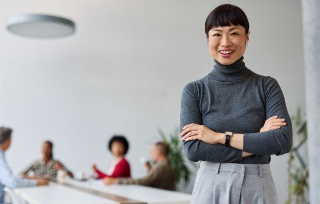 Vorgesetzte steht im Vordergrund und l&auml;chelt in die Kamera und hinter ihr sitzt das Team
