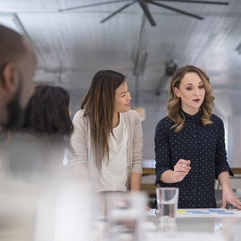 Weiterbildung Organisation, Personal, Führung Mehrere Personen stehen und sitzen um einen Tisch. Eine Frau ist im Fokus und spricht. Die anderen Personen hören ihr zu und gucken sie an.
