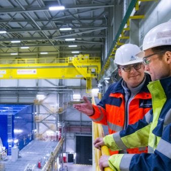 zu sehen zwei Männer mit Arbeitsschutzausrüstung und Blick in die Industriehalle