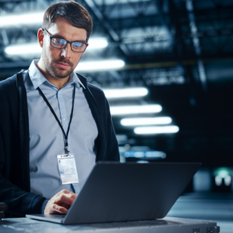 Junger Mann mit Brille steht in einer Halle vor einem aufgeklappten Laptop