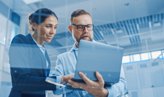 Eine Frau und ein Mann stehen gemeinsam und schauen auf einen Laptop, den der Mann in der Hand hält. Die Umgebung ist in einem Büro und wirkt modern.