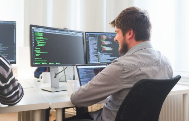 Ein Mann sitzt an einem Schreibtisch in einem modernen B&uuml;ro und arbeitet an einem Computer mit mehreren Bildschirmen. Auf den Monitoren sind Programmiercodes und Entwicklungsumgebungen zu sehen.