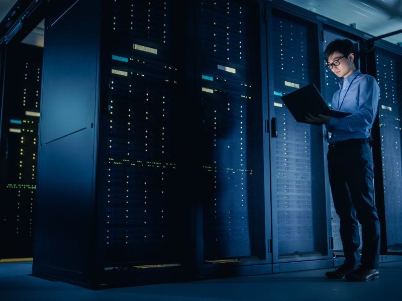 Ein Mann steht in einem Serverraum mit einem geöffneten laptop in der Hand