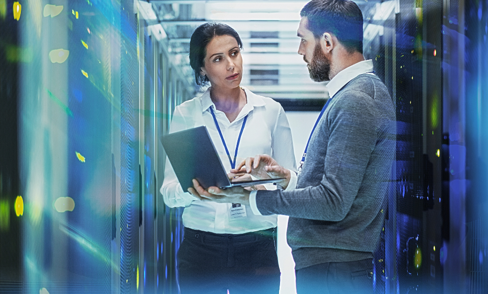 Ein Mann und eine Frau unterhalten sich im Serverraum und haben einen Laptop in der Hand