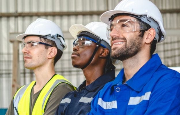 Drei Industriearbeiter in Schutzhelmen und Arbeitskleidung stehen in einer Halle und blicken nach oben.