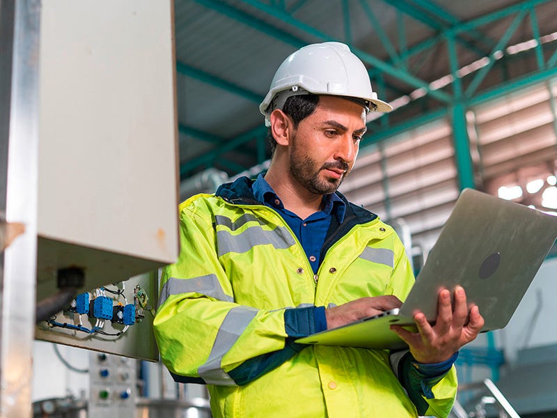 Ein Mann mit Sicherheitshelm und Arbeitsjacke hält einen Laptop in einer Produktionshalle in der Hand
