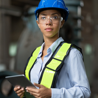 Eine Frau tr&auml;gt in einer industriellen Umgebung einen blauen Schutzhelm, eine Schutzbrille und eine gelbe Warnweste. Sie h&auml;lt ein Tablet in den H&auml;nden und blickt direkt in die Kamera.