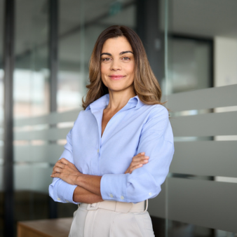 Eine Frau steht in einem modernen B&uuml;ro mit verschr&auml;nkten Armen vor einer Glaswand. Sie tr&auml;gt eine hellblaue Bluse und eine helle Hose und blickt mit leichtem L&auml;cheln in die Kamera.