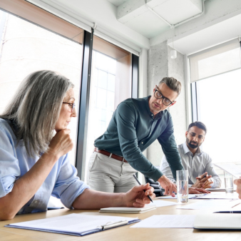 F&uuml;nf Personen sitzen in einem hellen Besprechungsraum um einen Tisch. Ein Mann steht leicht nach vorn gebeugt und zeigt auf Unterlagen, w&auml;hrend die anderen aufmerksam zuh&ouml;ren.