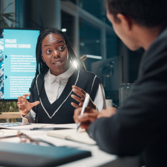 Drei Kolleginnen sitzen in einem modernen B&uuml;ro an einem Besprechungstisch. Eine Frau gestikuliert beim Sprechen, w&auml;hrend im Hintergrund auf einem Bildschirm eine Pr&auml;sentation zum Thema maschinelles Lernen zu sehen ist.