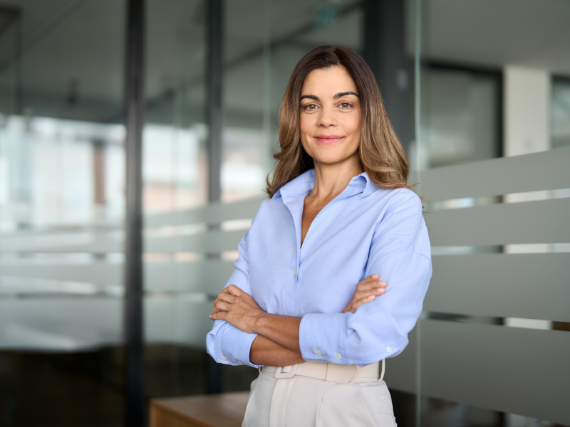 Eine Frau steht in einem modernen B&uuml;ro mit verschr&auml;nkten Armen vor einer Glaswand. Sie tr&auml;gt eine hellblaue Bluse und eine helle Hose und blickt mit leichtem L&auml;cheln in die Kamera.