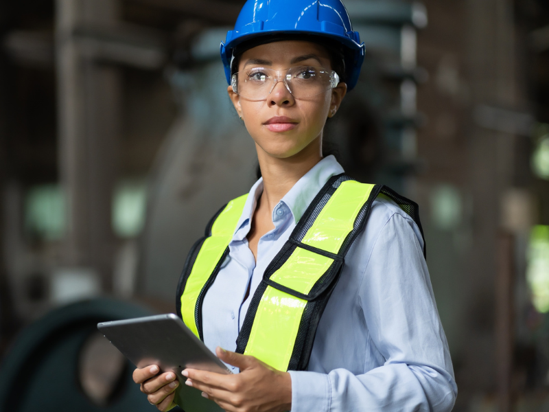 Eine Frau tr&auml;gt in einer industriellen Umgebung einen blauen Schutzhelm, eine Schutzbrille und eine gelbe Warnweste. Sie h&auml;lt ein Tablet in den H&auml;nden und blickt direkt in die Kamera.