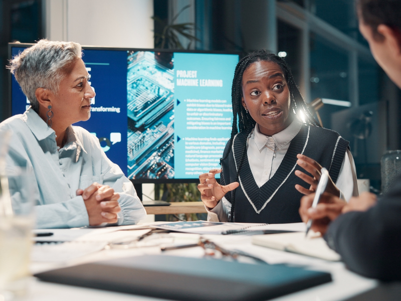 Drei Kolleginnen sitzen in einem modernen B&uuml;ro an einem Besprechungstisch. Eine Frau gestikuliert beim Sprechen, w&auml;hrend im Hintergrund auf einem Bildschirm eine Pr&auml;sentation zum Thema maschinelles Lernen zu sehen ist.