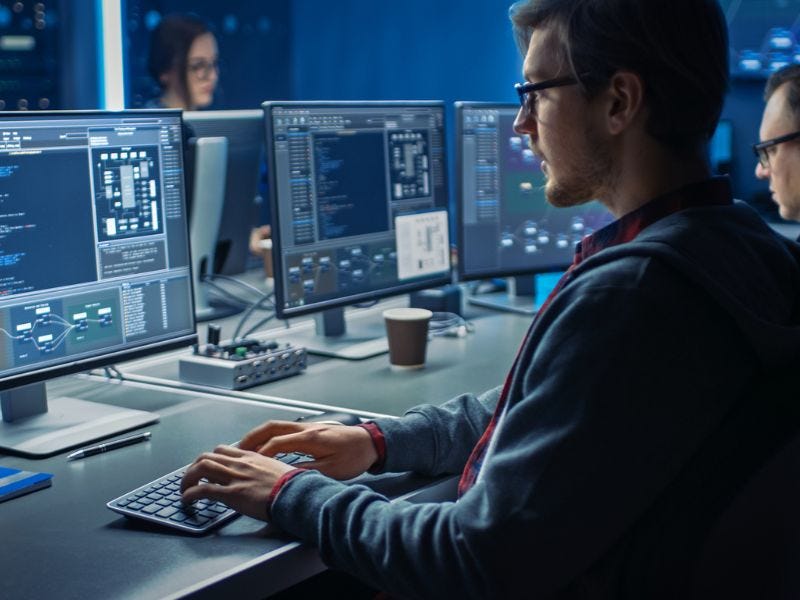 Mann mit Brille sitzt am Computer. Vier Bildschirme mit vielen Daten sind zu sehen. Zwei weitere Personen sind im Hintergrund.