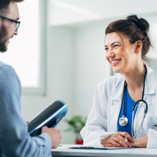 Lächelende Frau im Arztkittel und mit umhängendem Stethoskop führt ein Gespräch mit einem ihr gegenübersitzenden Mann.