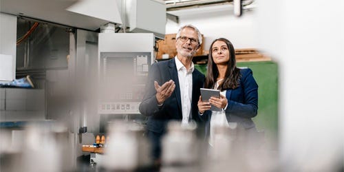 Zwei Qualit&auml;tsmanager besprechen sich in einer industriellen Produktionsumgebung und pr&uuml;fen gemeinsam Unterlagen auf einem Tablet.