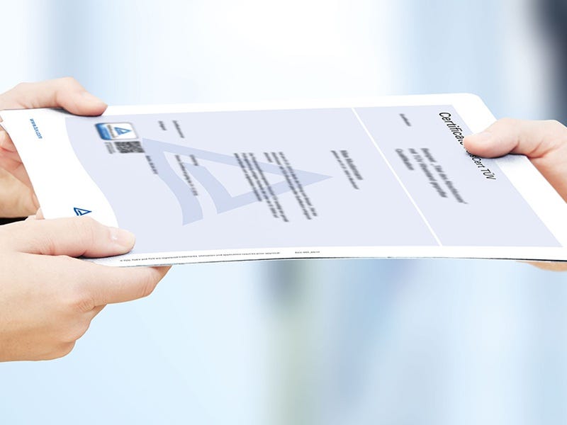 Zwei H&auml;nde &uuml;berreichen ein Zertifikat mit T&Uuml;V- Logo vor unscharfem, hellem Hintergrund.