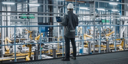 Person mit Schutzhelm &uuml;berwacht eine moderne Produktionshalle mit Robotikarmen und digitalem Steuerungssystem.
