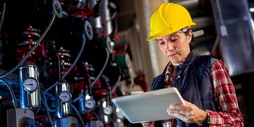 Ingenieurin mit gelbem Schutzhelm überprüft technische Anlagen mithilfe eines Tablets in einer Industrieumgebung.