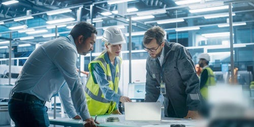Drei Fachkr&auml;fte stehen zusammen an einem Laptop und besprechen Arbeitsabl&auml;ufe in einer industriellen Umgebung.