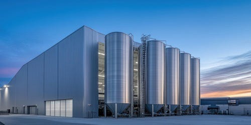 Moderne Industriehalle mit f&uuml;nf gro&szlig;en Edelstahl-Silos im Au&szlig;enbereich bei Abendlicht und klarem Himmel.
