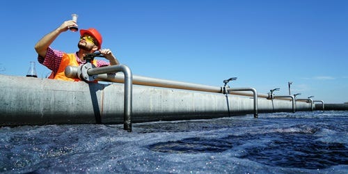 Techniker mit Helm und Warnweste untersucht eine Flüssigkeitsprobe an einer großen Wasseraufbereitungsanlage.