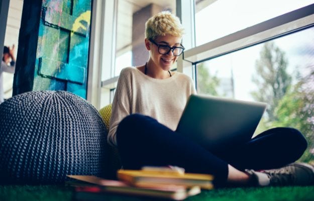 Junge Frau mit kurzen Haaren und Brille sitzt am Notebook im Schneidersitz